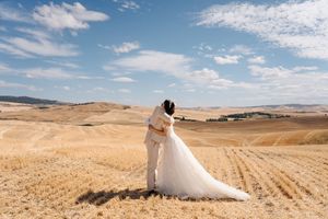 A wedding dress train flows across a golden wheat field under bright blue skies with scattered white clouds.