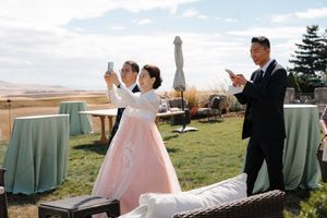 A wedding celebration takes place outdoors on a grassy hilltop with light blue chairs and pink decor accents.