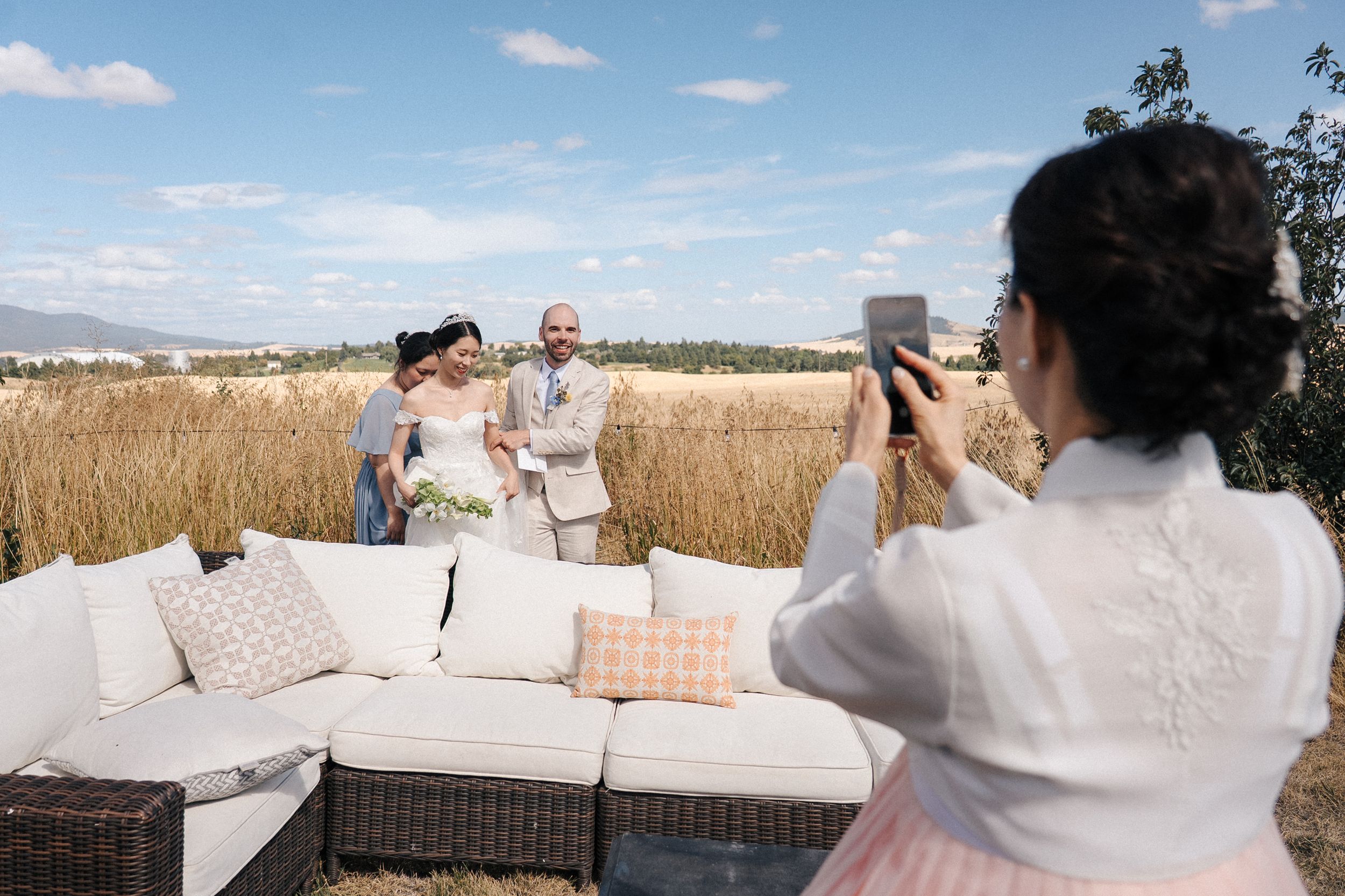 A photographer captures a moment at an outdoor lounge area with white cushions against a backdrop of golden wheat fields.