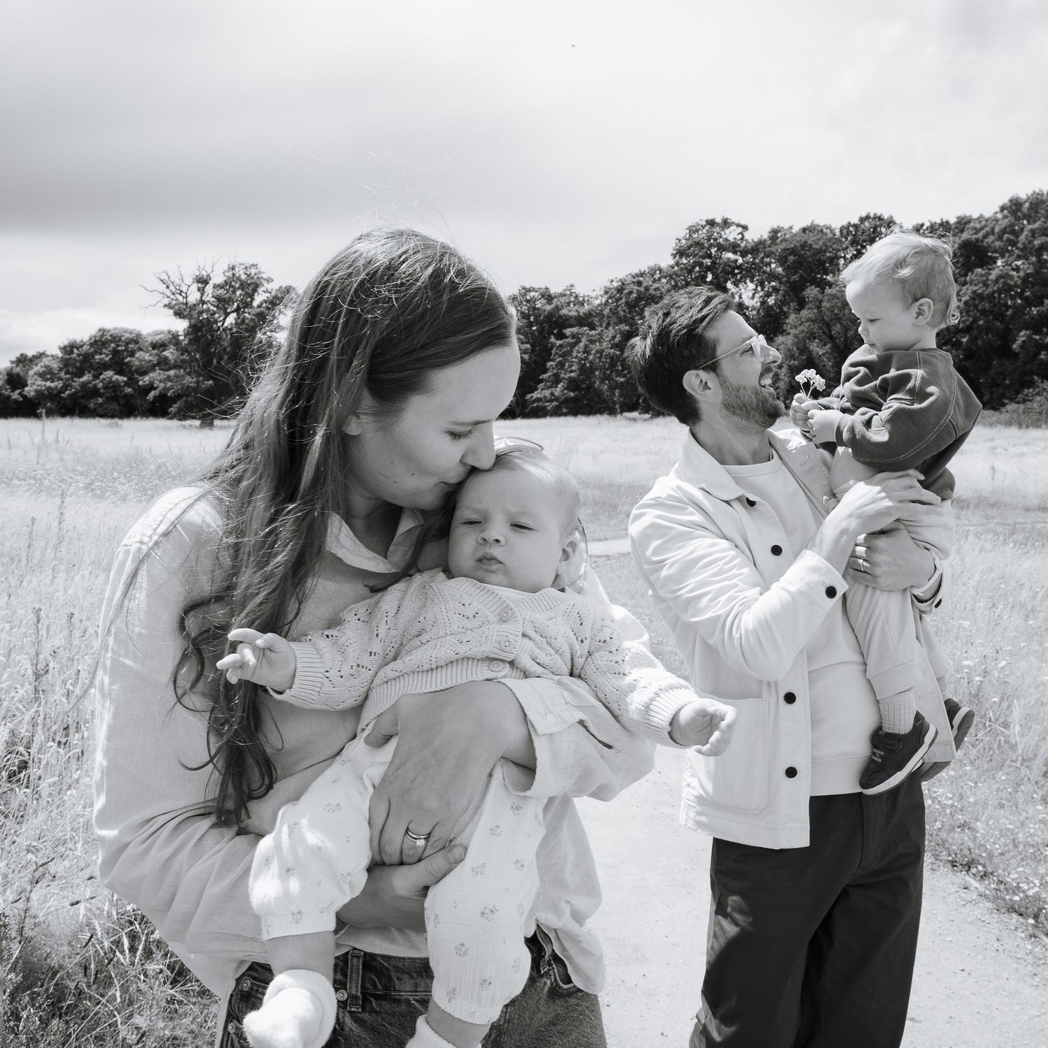 Black and white photo of a Mum kissing her baby daughter. Dad is just behind holding his older sun.  Unposed photo of the family in a meadow