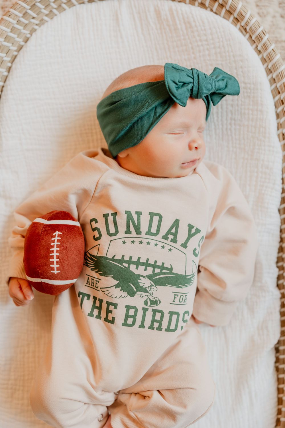 Sleeping newborn baby lies in a basket wearing a green headband and holding a small football.