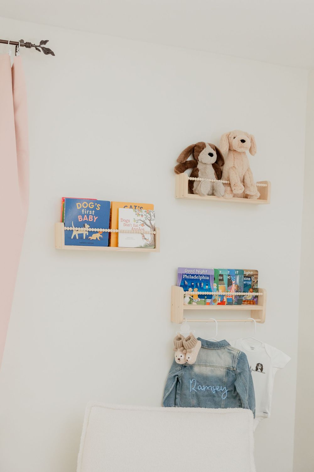Wooden nursery shelves hold baby books, stuffed dogs, and a small denim jacket hanging on the wall.