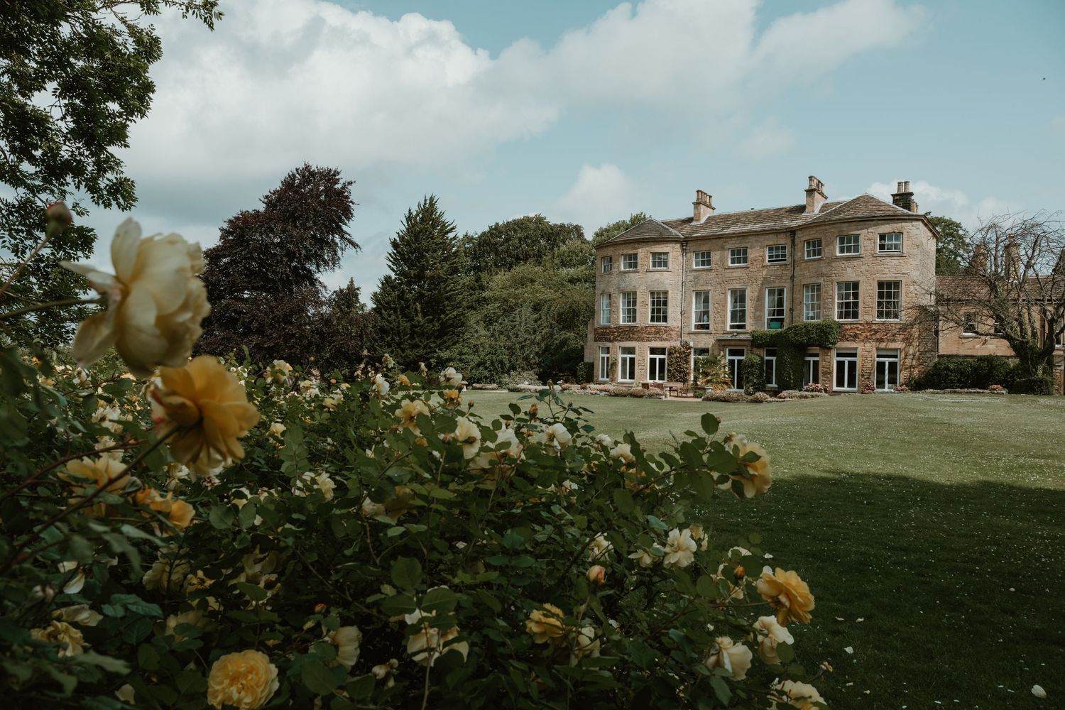 Elegant Georgian manor house with yellow roses blooming in the foreground and manicured lawn on a sunny day.