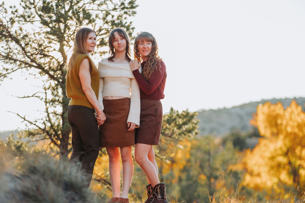 Three people in brown and white outfits pose together against a scenic mountain backdrop during golden hour.