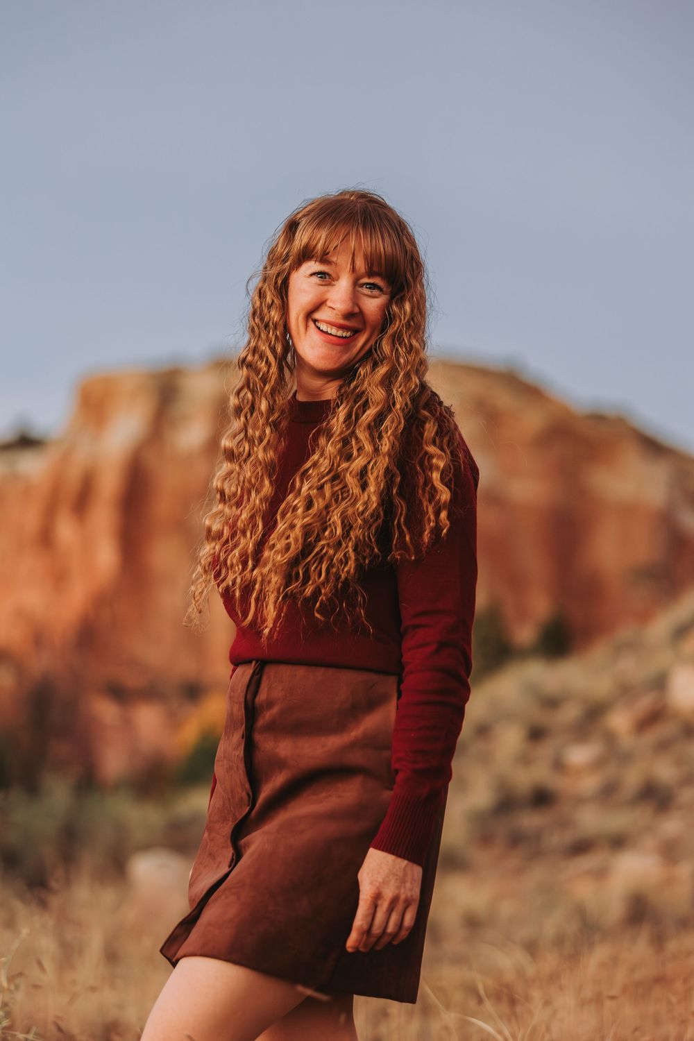 A person with curly red hair wearing a brown sweater poses against a backdrop of red rock formations in a desert landscape.