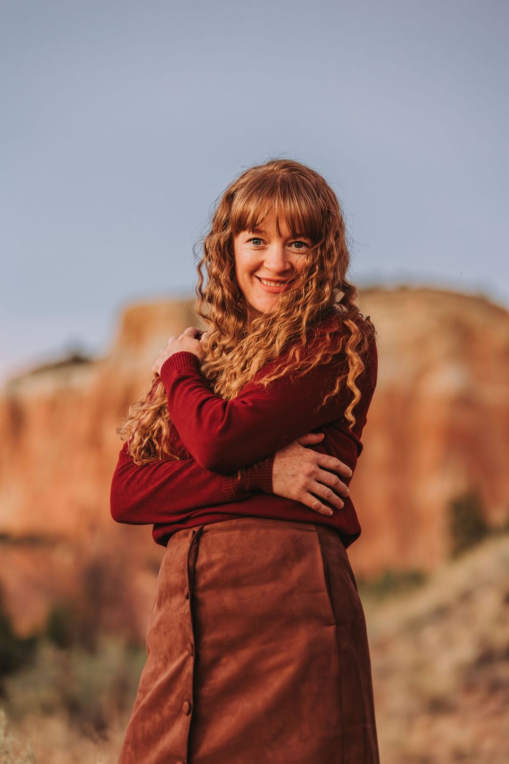 Person in a burgundy sweater hugging themselves against a backdrop of red rock formations in warm sunlight.