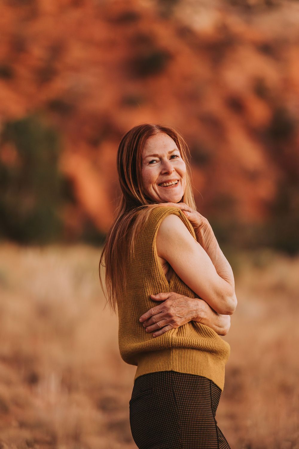 Person in a brown knit sweater embracing themselves with joy against desert rock formations at sunset.