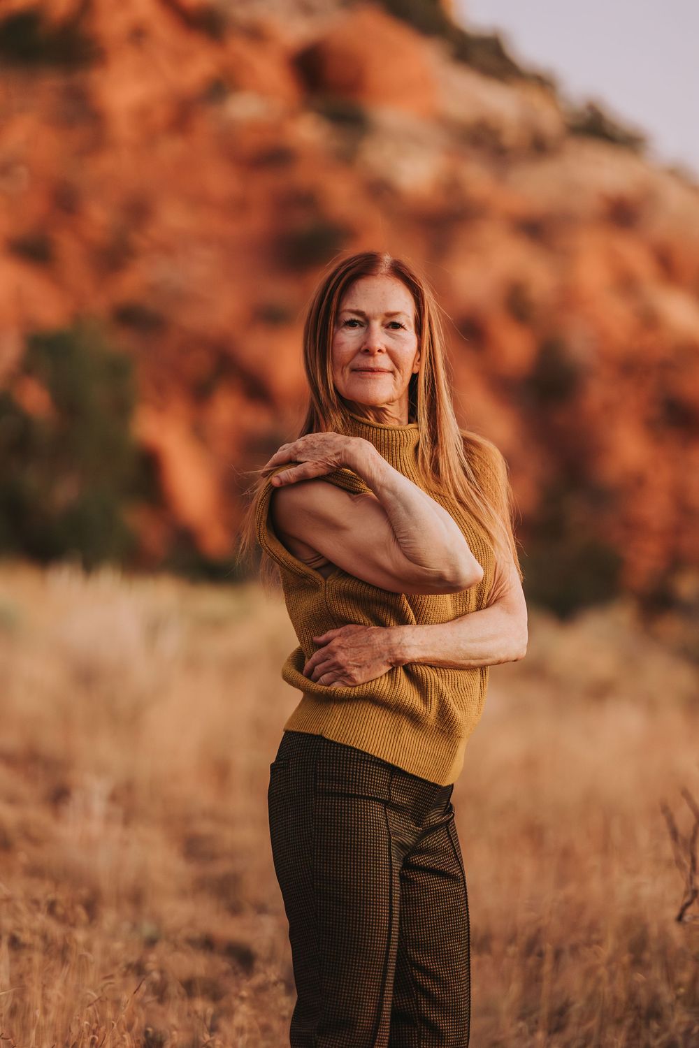 Person in earth-toned clothing stands against backdrop of red desert rocks during golden hour.