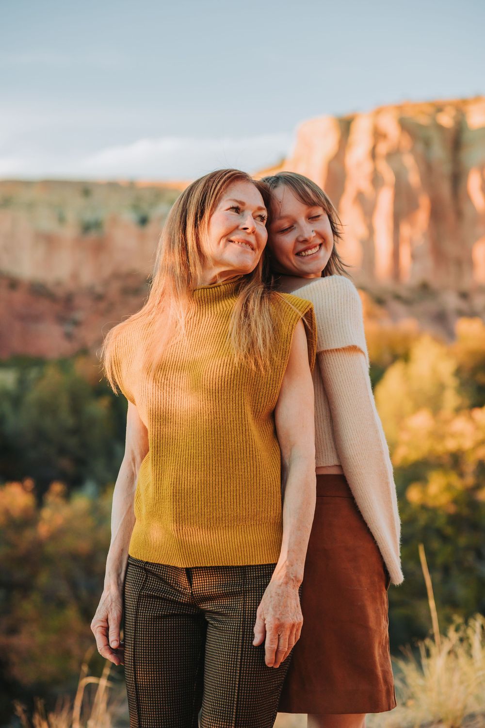 Two friends in warm autumn colors share a joyful moment against a scenic desert canyon backdrop at sunset.