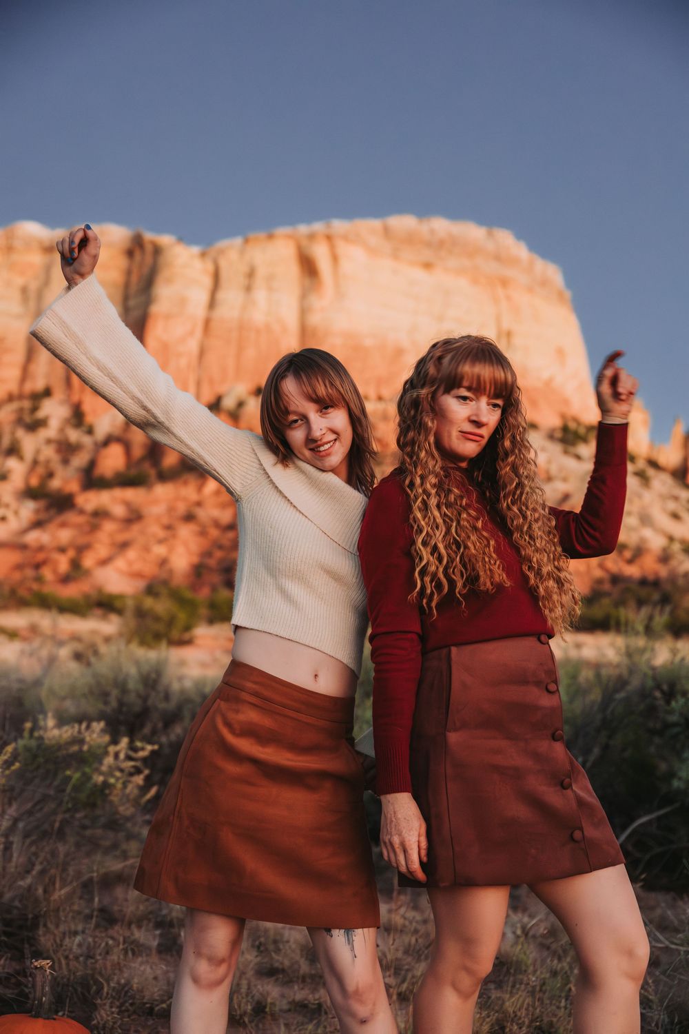 Two women in autumn-colored outfits pose playfully against red rock formations in a desert landscape at sunset.