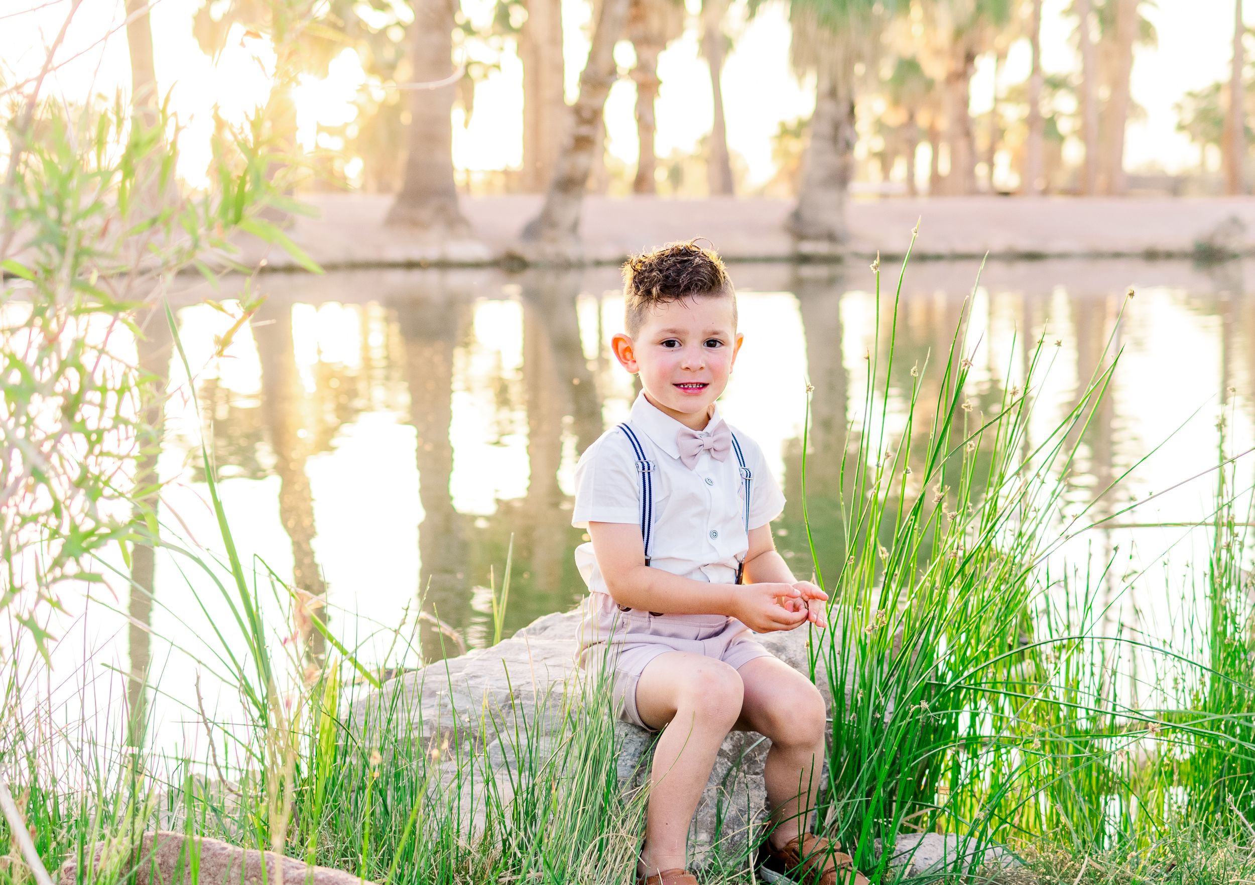 Light and airy portrait of a young toddler boy sitting on a rock in the middle of tall grass in front of a pond at Papago Park in Phoenix.
