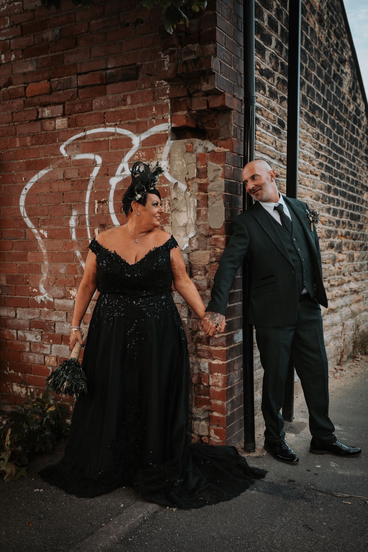 A couple in formal black attire poses against a rustic brick wall in an artistic photo series.