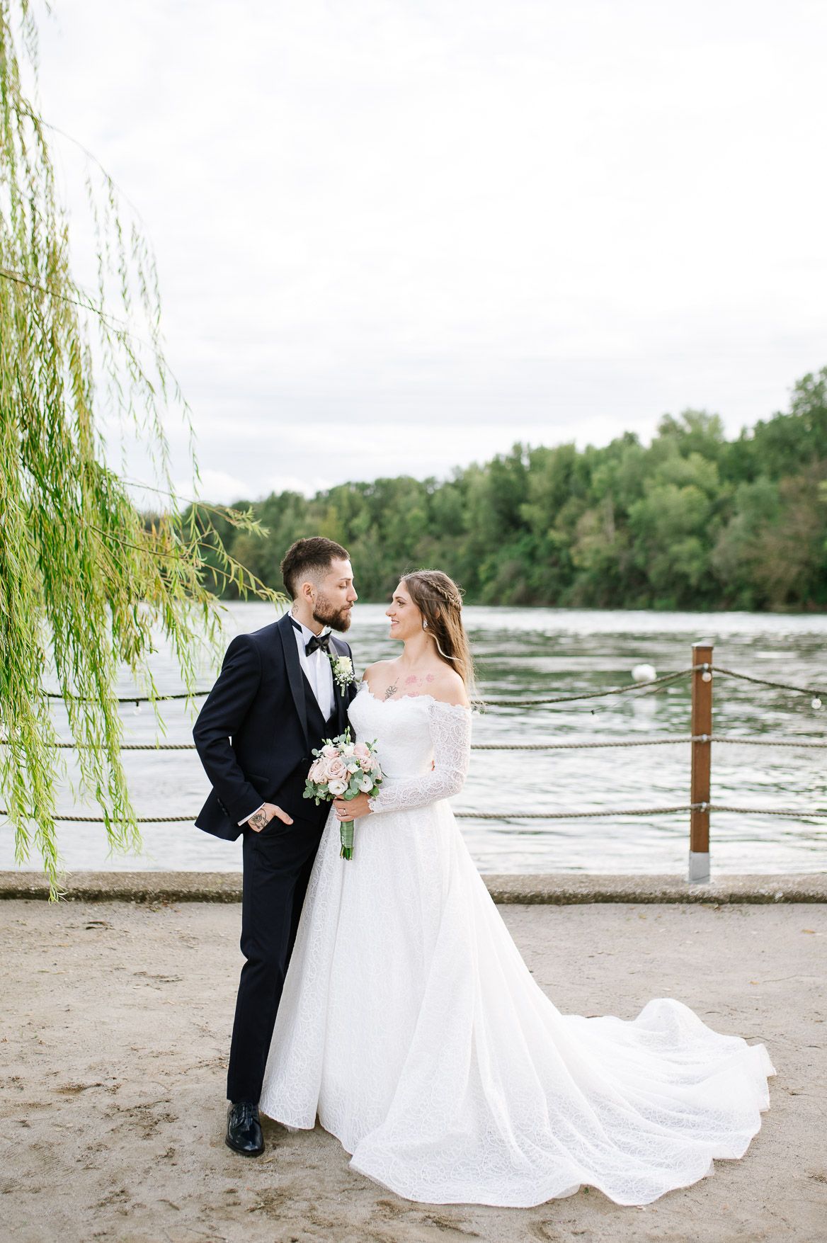 A couple in wedding attire stands by a riverbank with willow trees and water in the background.