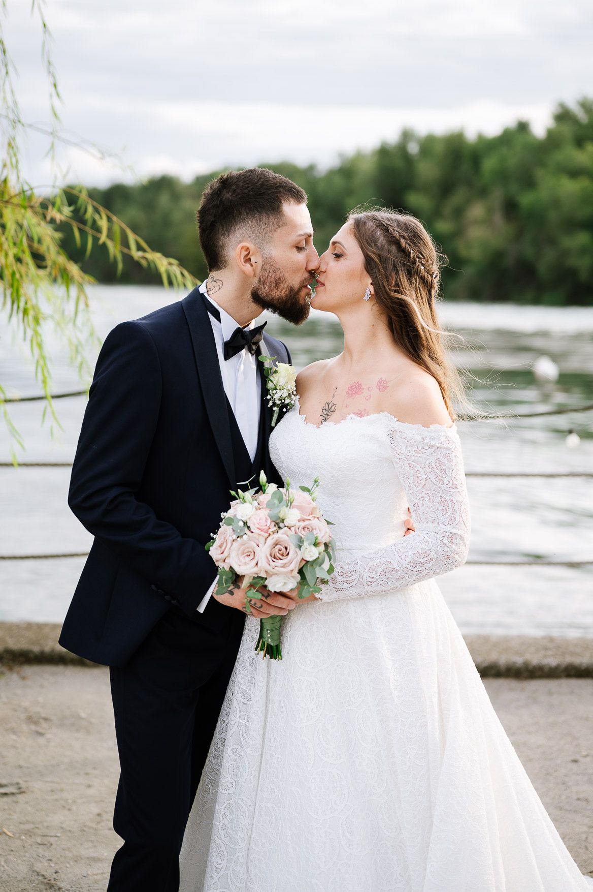 A romantic wedding couple shares intimate kisses by a lakeside with willow trees and pink floral bouquet.