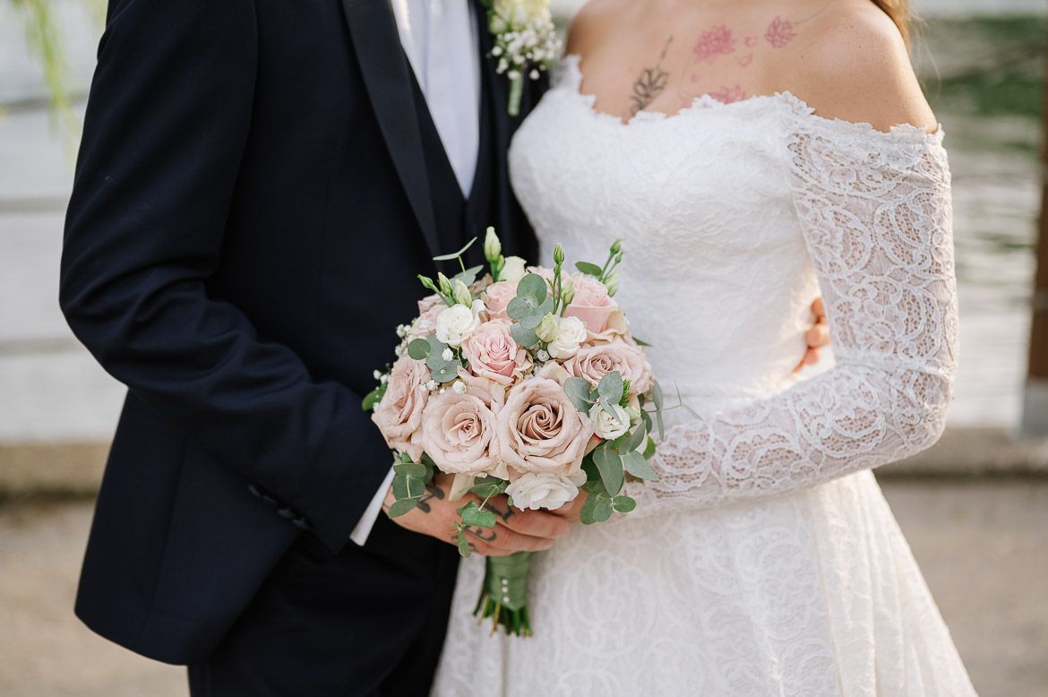 A wedding couple holds hands while holding a soft pink rose bouquet with white accents and greenery.