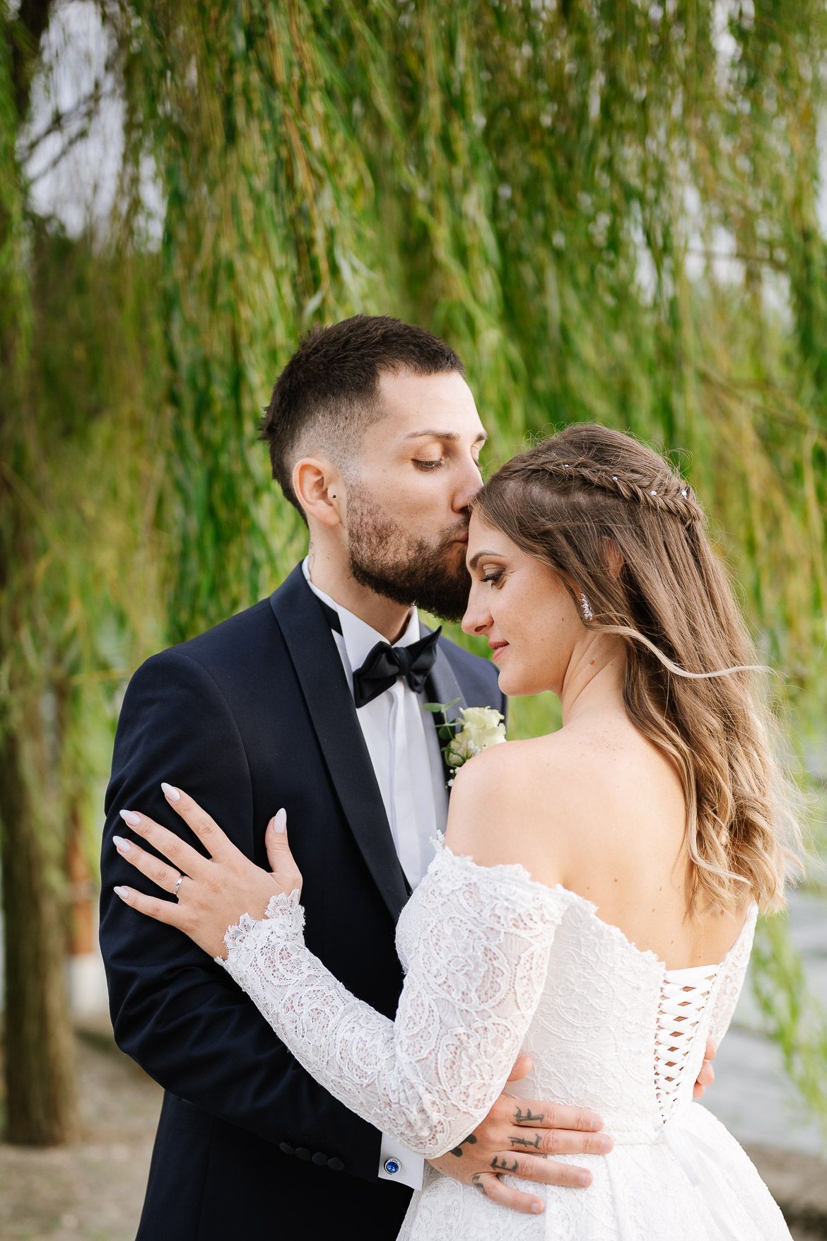 A romantic wedding couple embraces under cascading willow trees in an intimate outdoor setting.