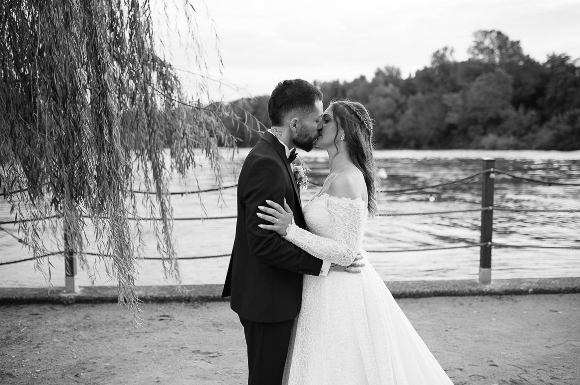 A couple in formal wedding attire embraces by a lake with willow trees creating a romantic outdoor setting.