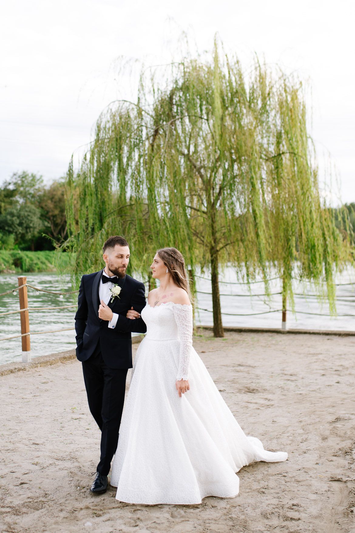A couple shares a romantic moment under willow trees near a lake on their wedding day.