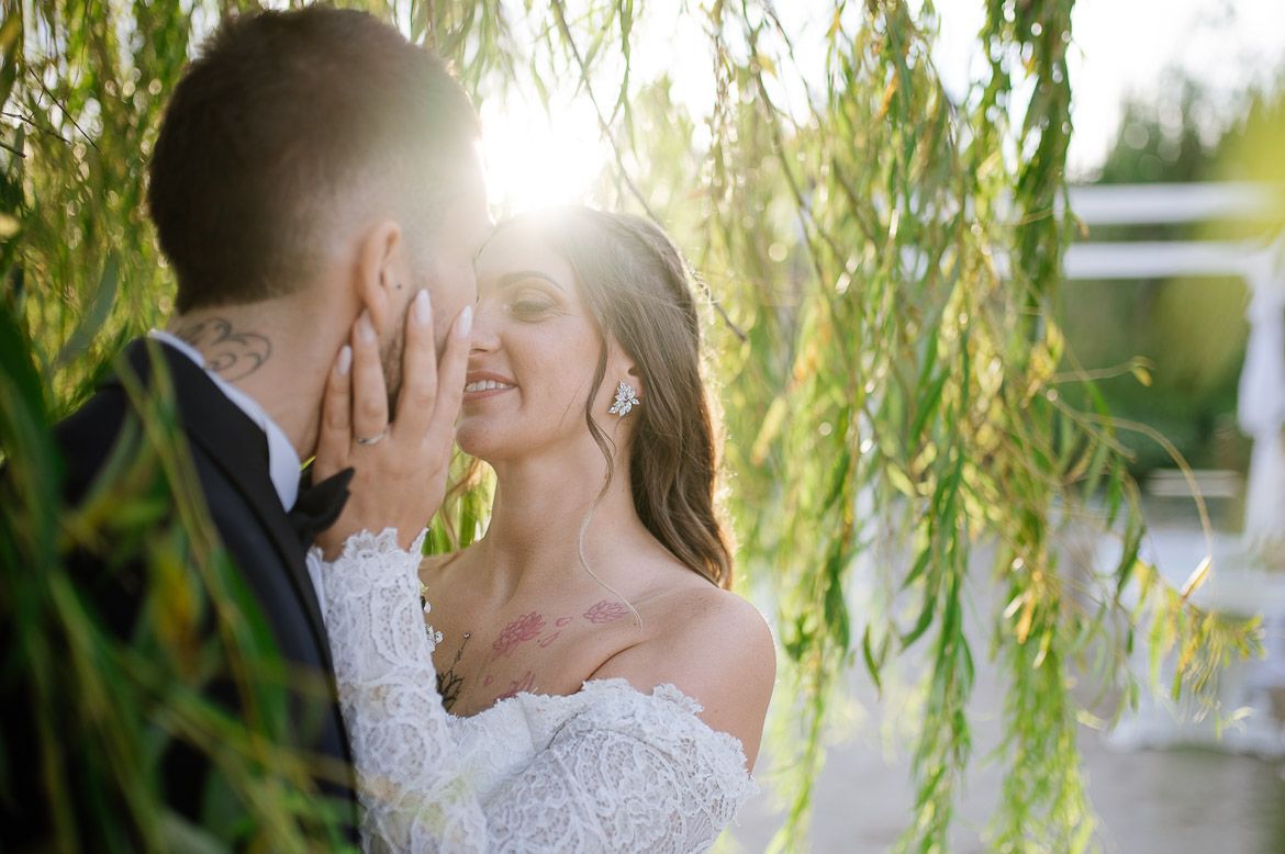 A romantic wedding moment captured through willow tree branches with sunlight creating a dreamy backlit glow.