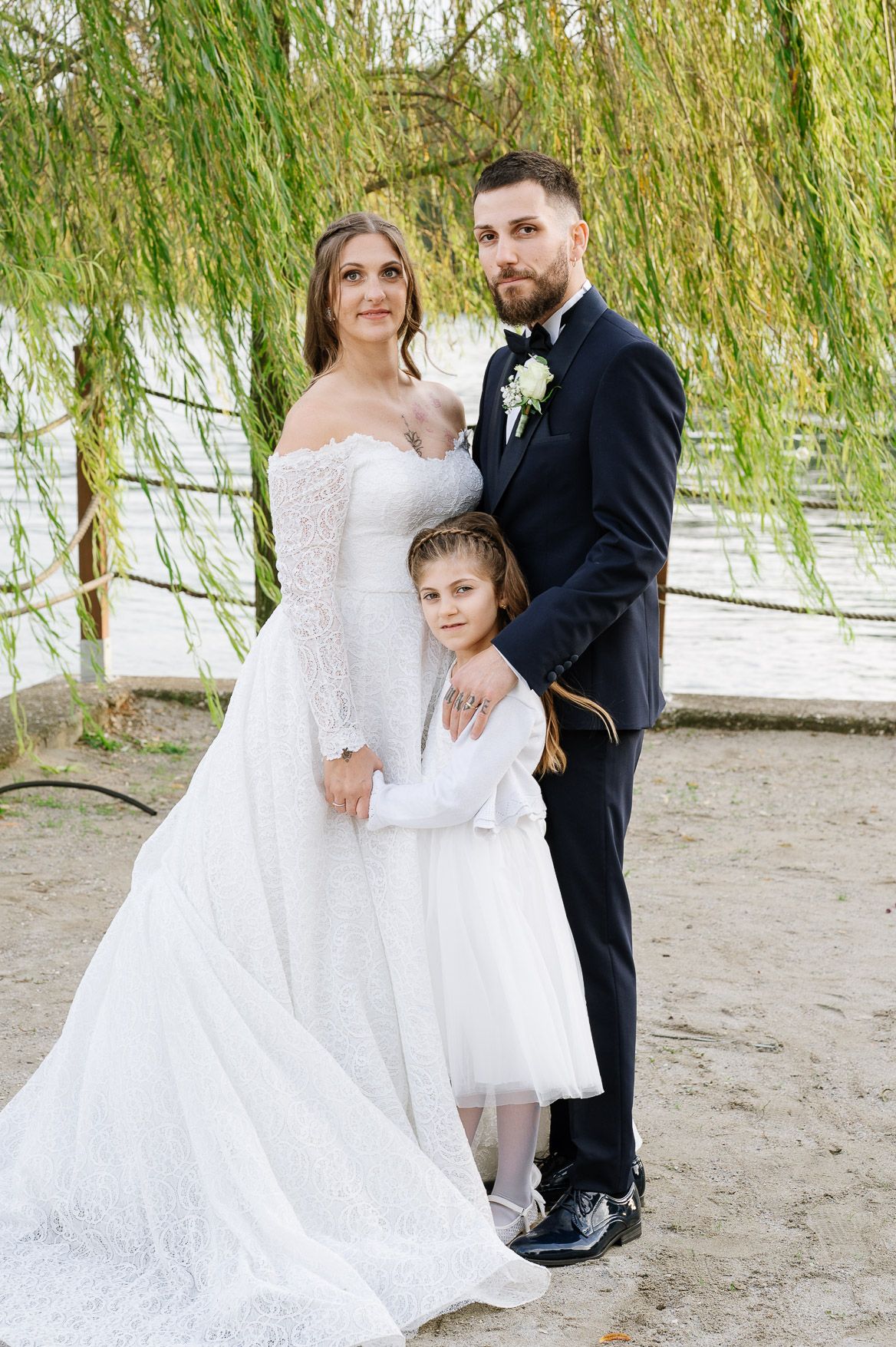 Wedding couple and child pose together under drooping willow branches near a sandy beach.