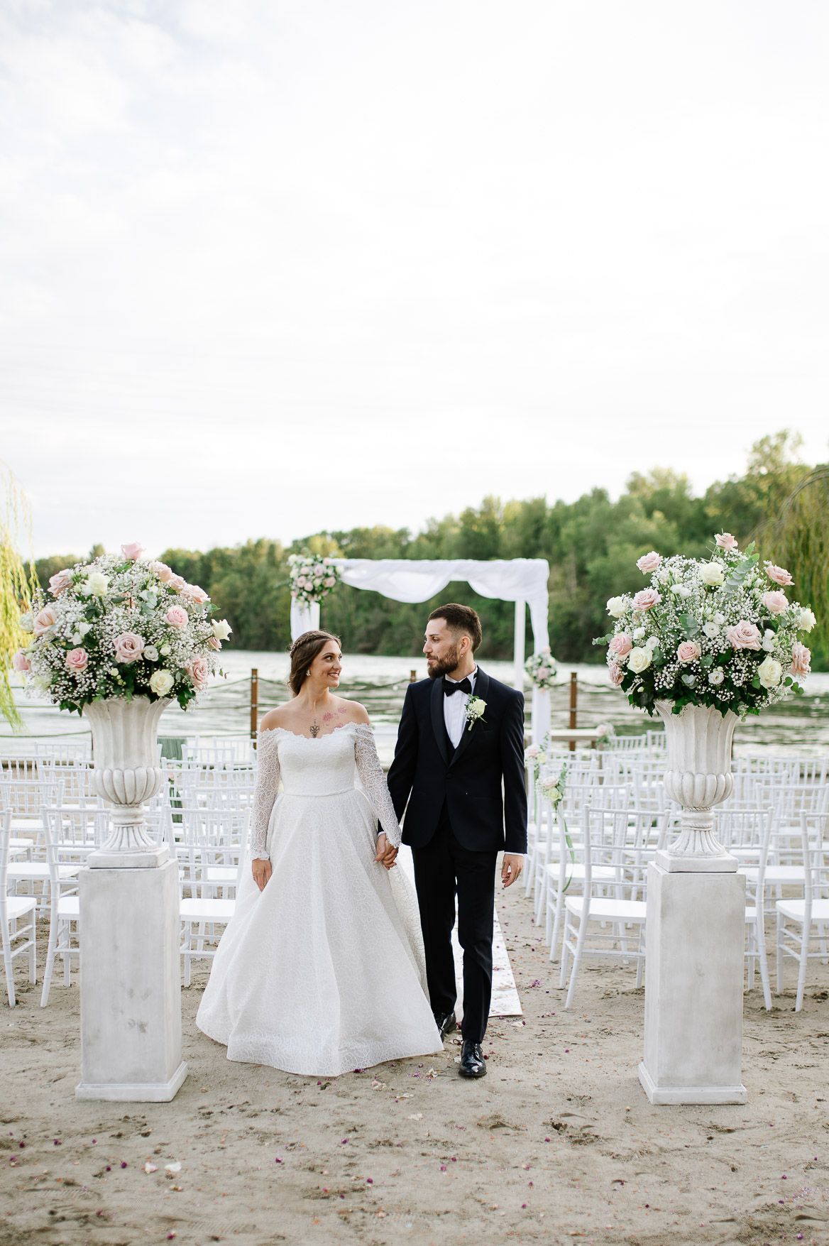 A romantic beach wedding ceremony setup with white chairs and elegant floral arrangements on white pedestals in the sand.