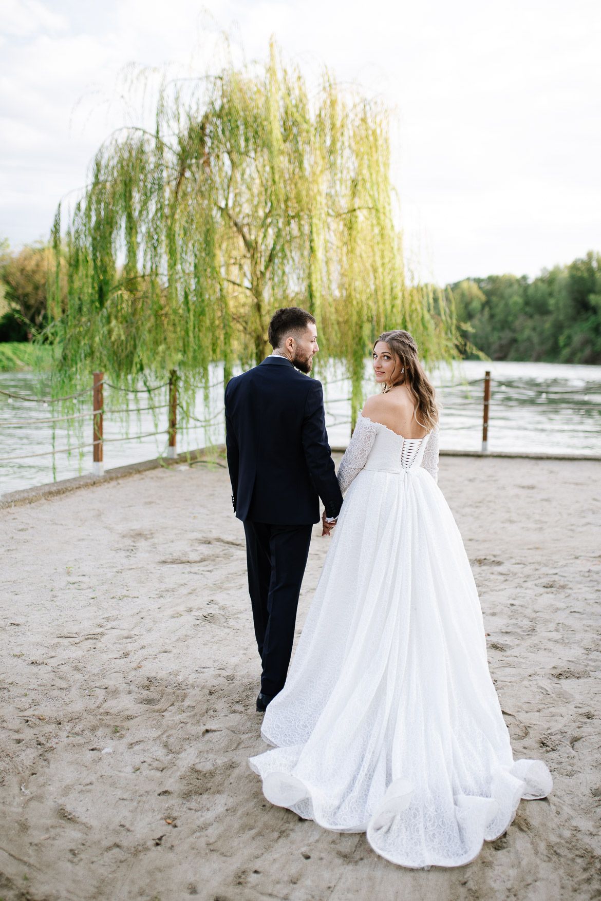 Newlyweds stroll along a sandy beach path beneath graceful weeping willow trees.