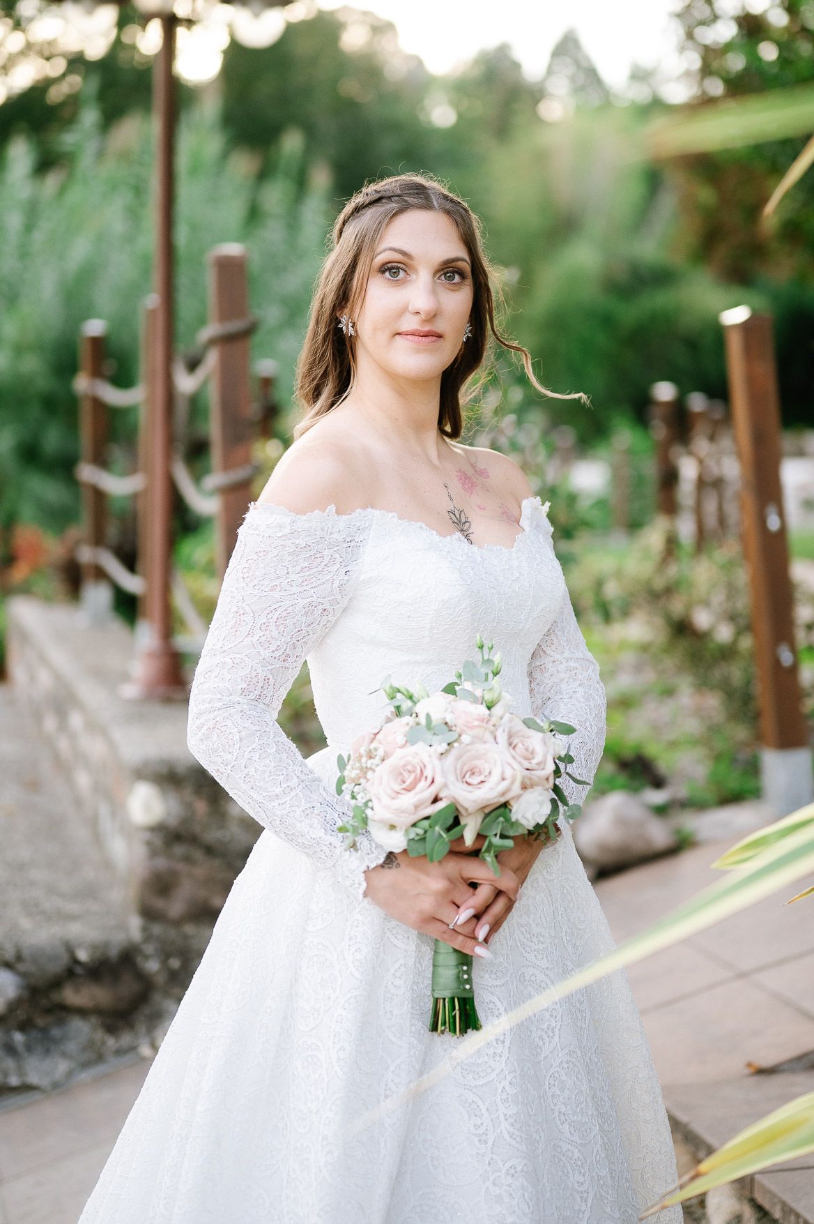 Bride in lace off-shoulder gown holds pink and white rose bouquet in outdoor garden setting.