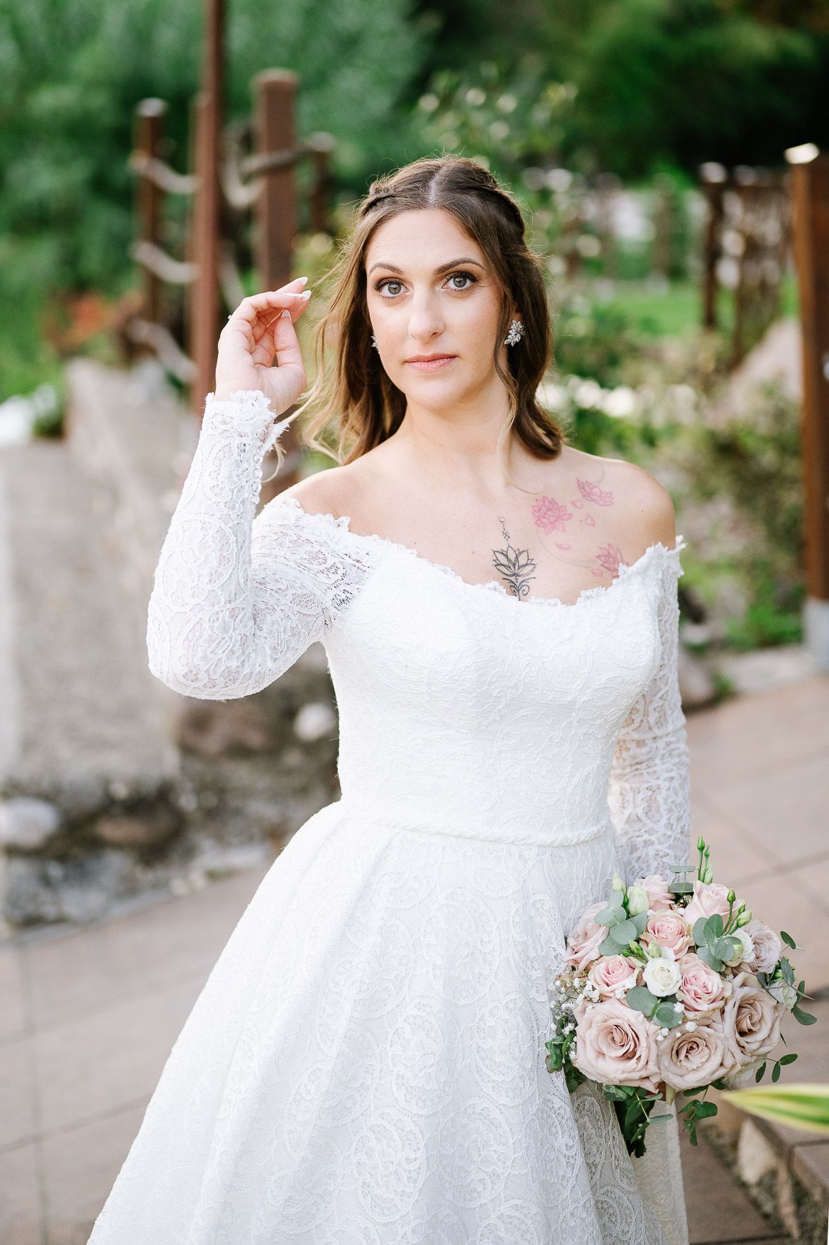 A bride in a long-sleeved lace wedding dress poses in a garden setting holding a bouquet of pink and white roses.