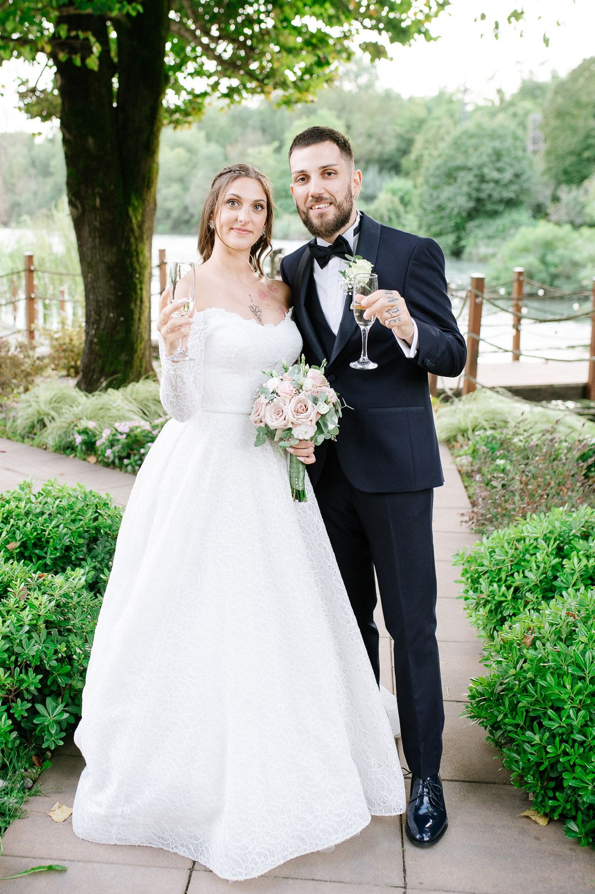 Elegant couple in wedding attire embraces on a landscaped walkway with blooming gardens.
