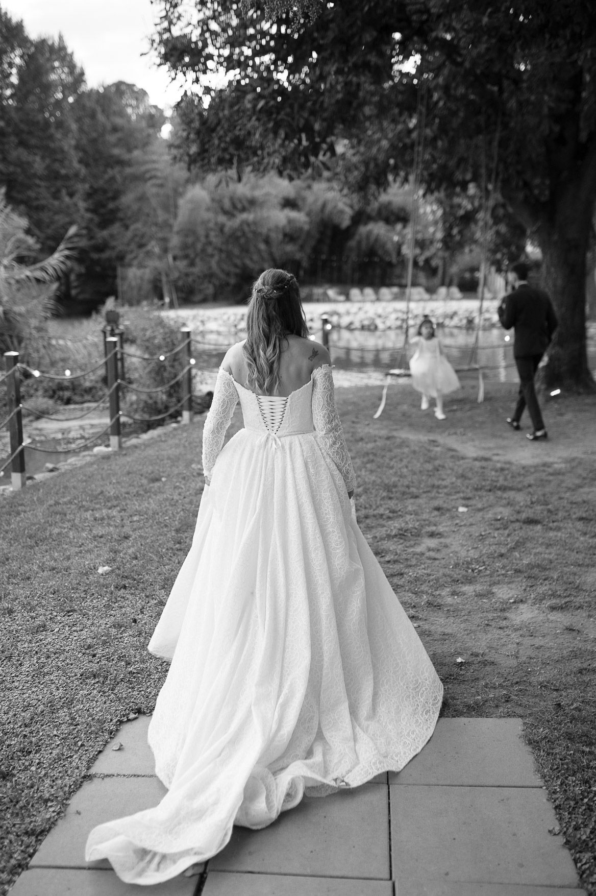 Black and white photograph of a flowing white wedding dress captured from behind on a garden path.