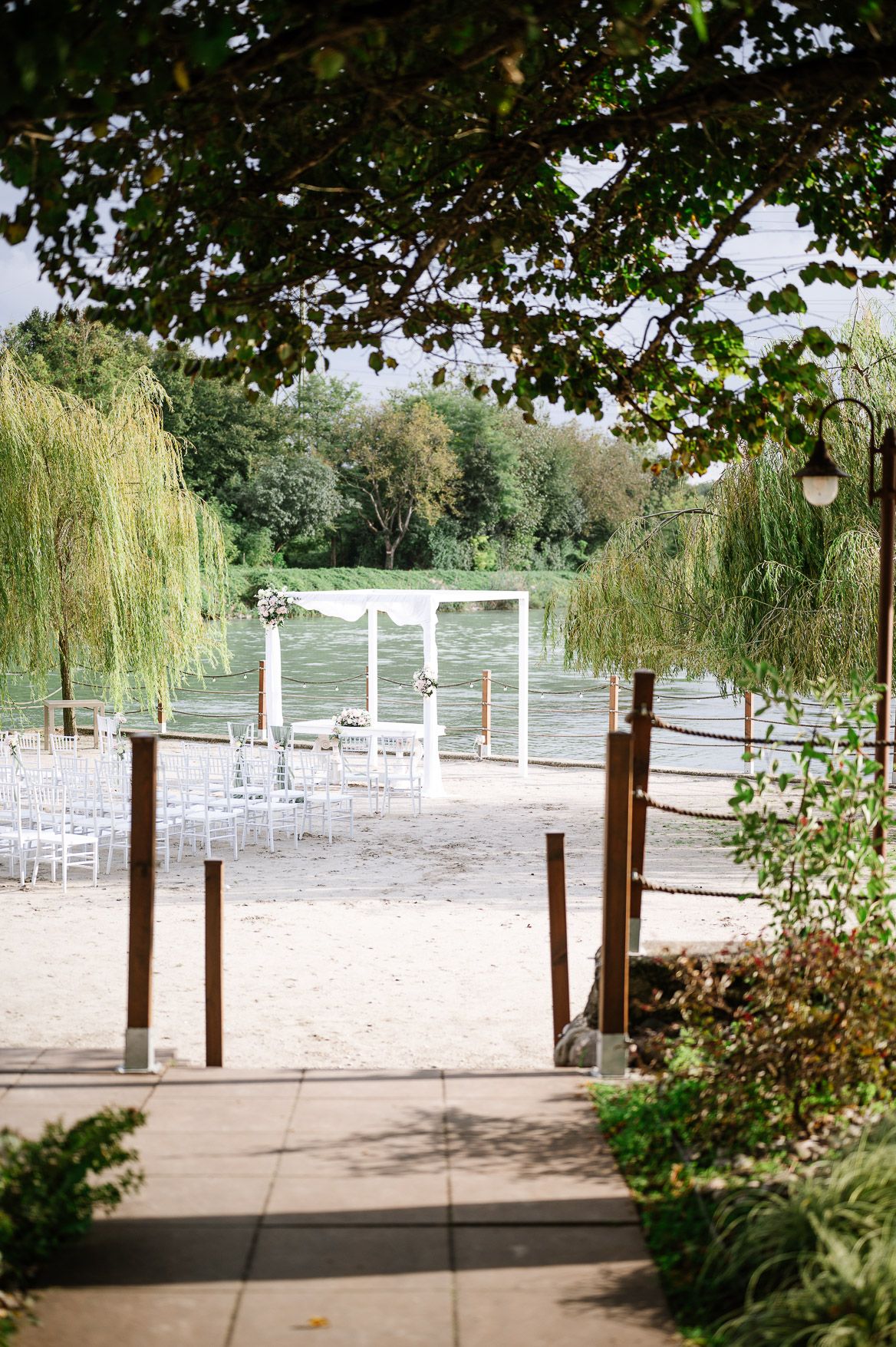 Outdoor wedding ceremony setup with white chairs arranged under trees near a waterfront.