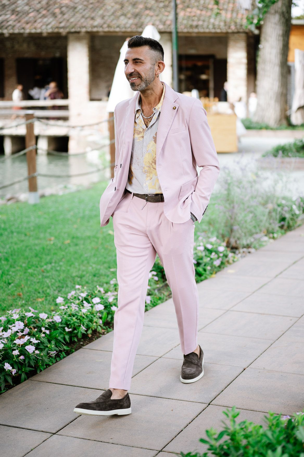 Man wearing a light pink suit and brown shoes walks along a stone pathway in a garden setting.