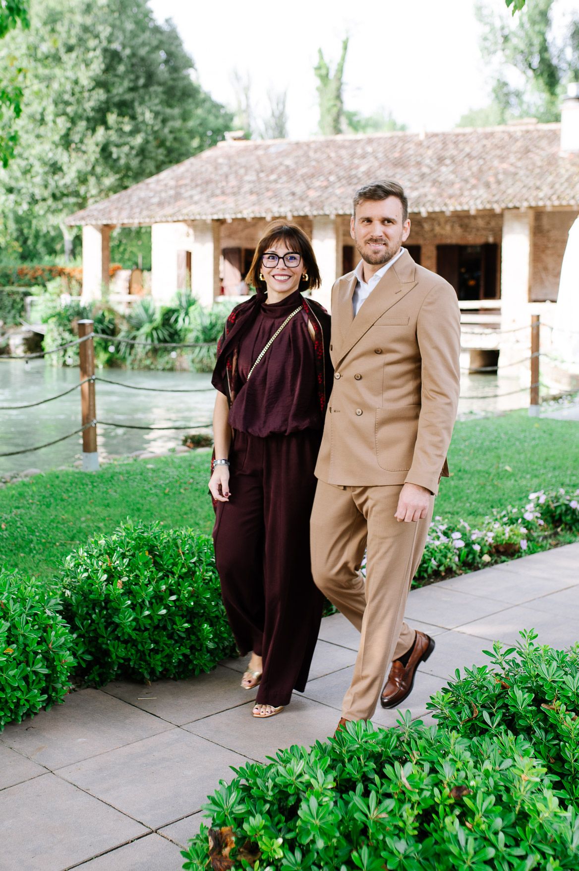 Two people in formal attire pose together on a garden pathway next to lush greenery and rustic buildings.