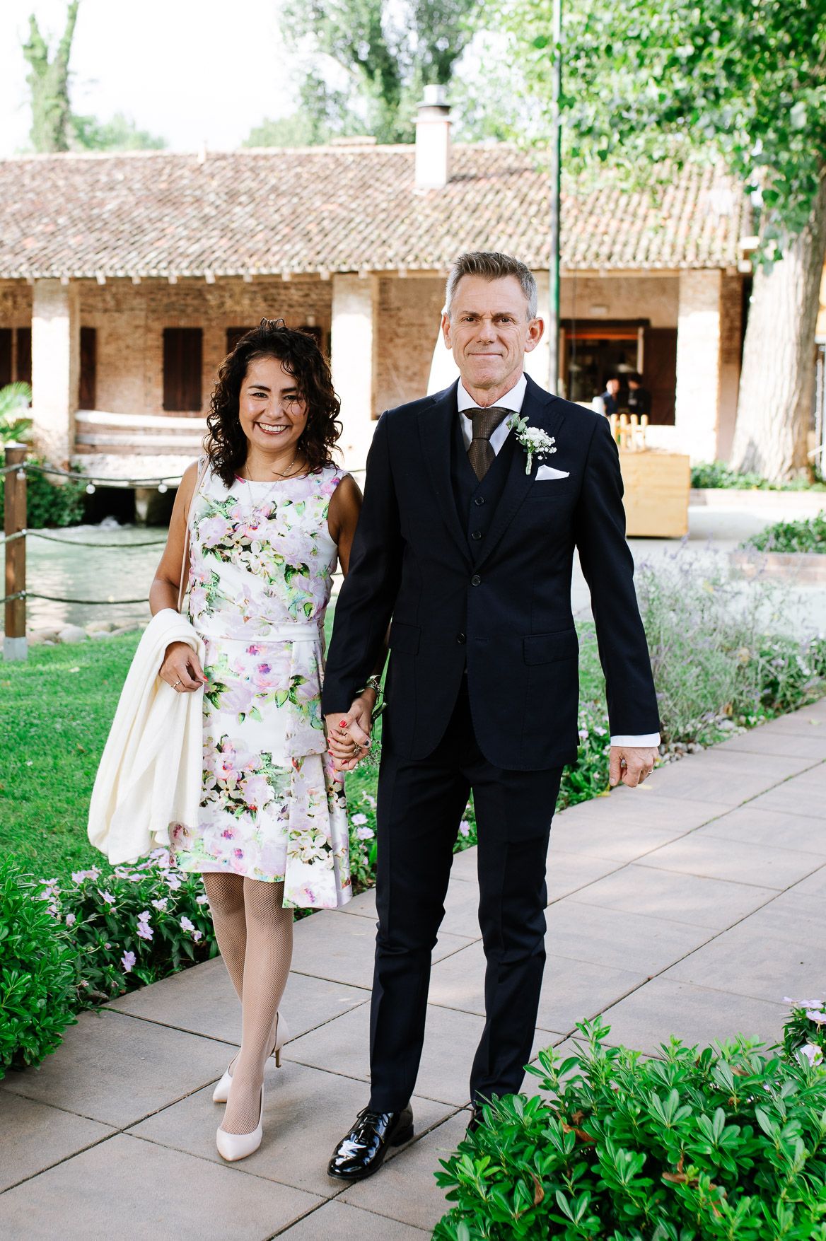 Wedding party members stroll along a garden pathway wearing formal black suits and floral dresses.