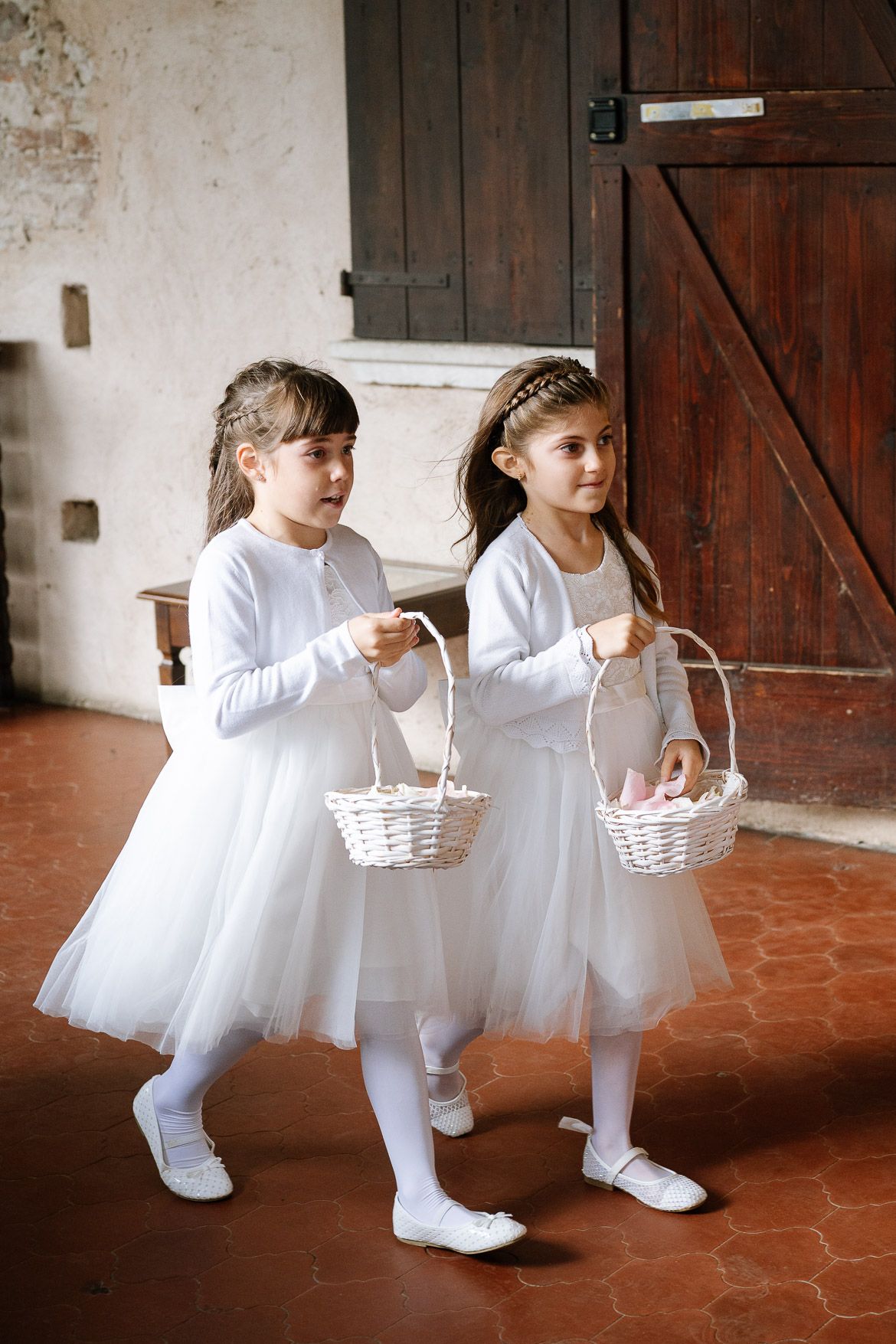Two flower girls in white dresses and tutus carry matching baskets down a rustic hallway.