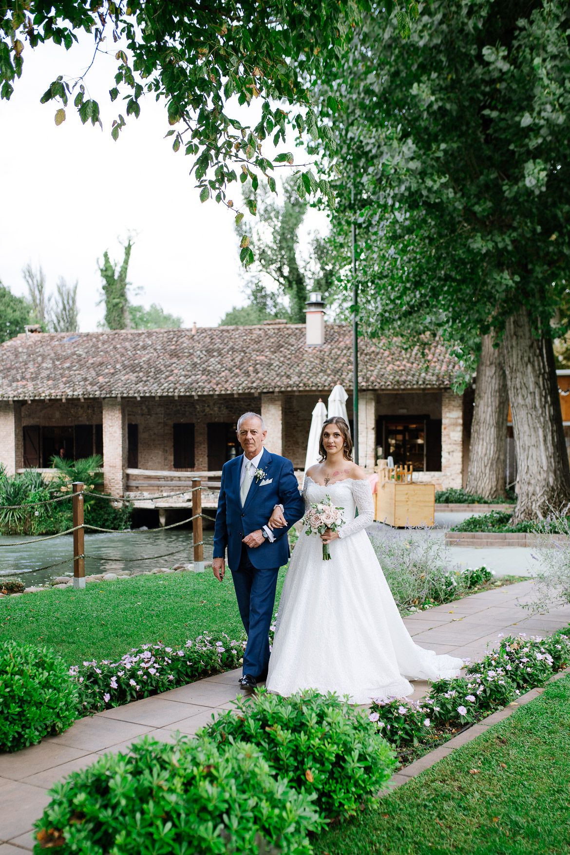 Couple walks down a scenic garden pathway lined with greenery and rustic adobe buildings.