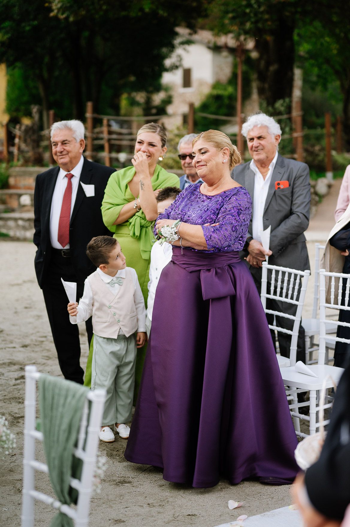 Wedding guests in formal attire gather around white chairs at an outdoor venue.