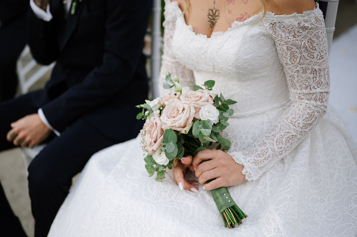 A delicate bridal bouquet of blush pink roses and greenery held against a white lace wedding dress.