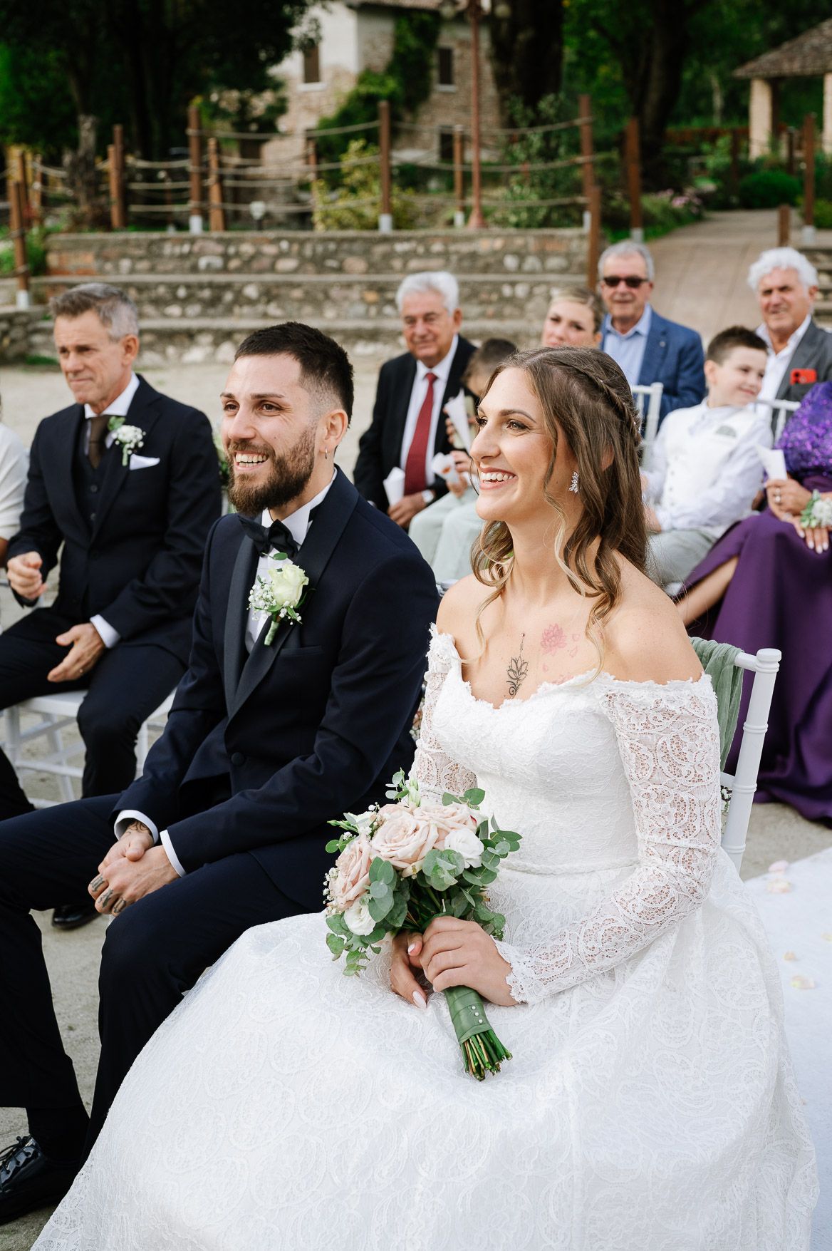 Happy couple laughs joyfully during outdoor wedding celebration while surrounded by guests in formal attire.