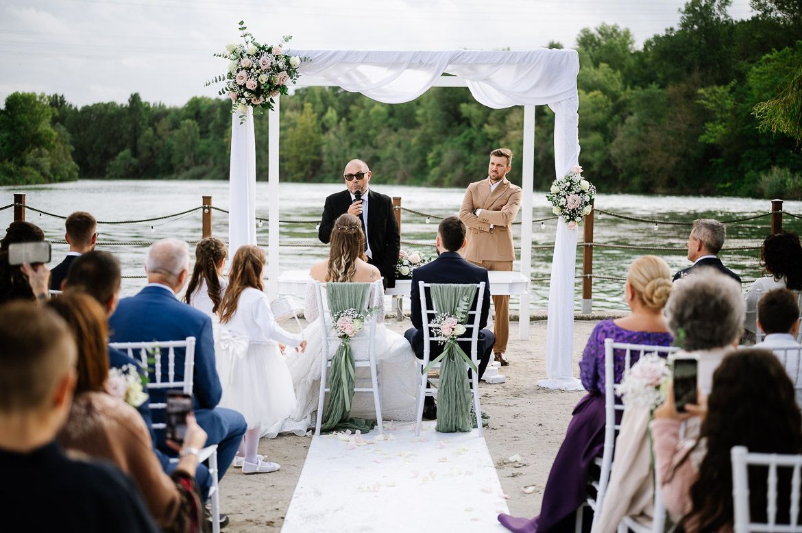 An outdoor wedding ceremony by a lake with white chairs and a floral arch on a cloudy day.