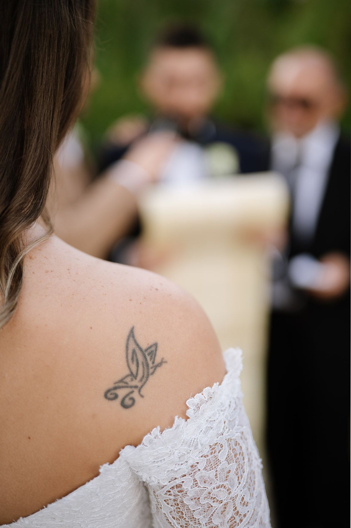 Close-up detail of a delicate lace wedding dress and small butterfly tattoo on shoulder.