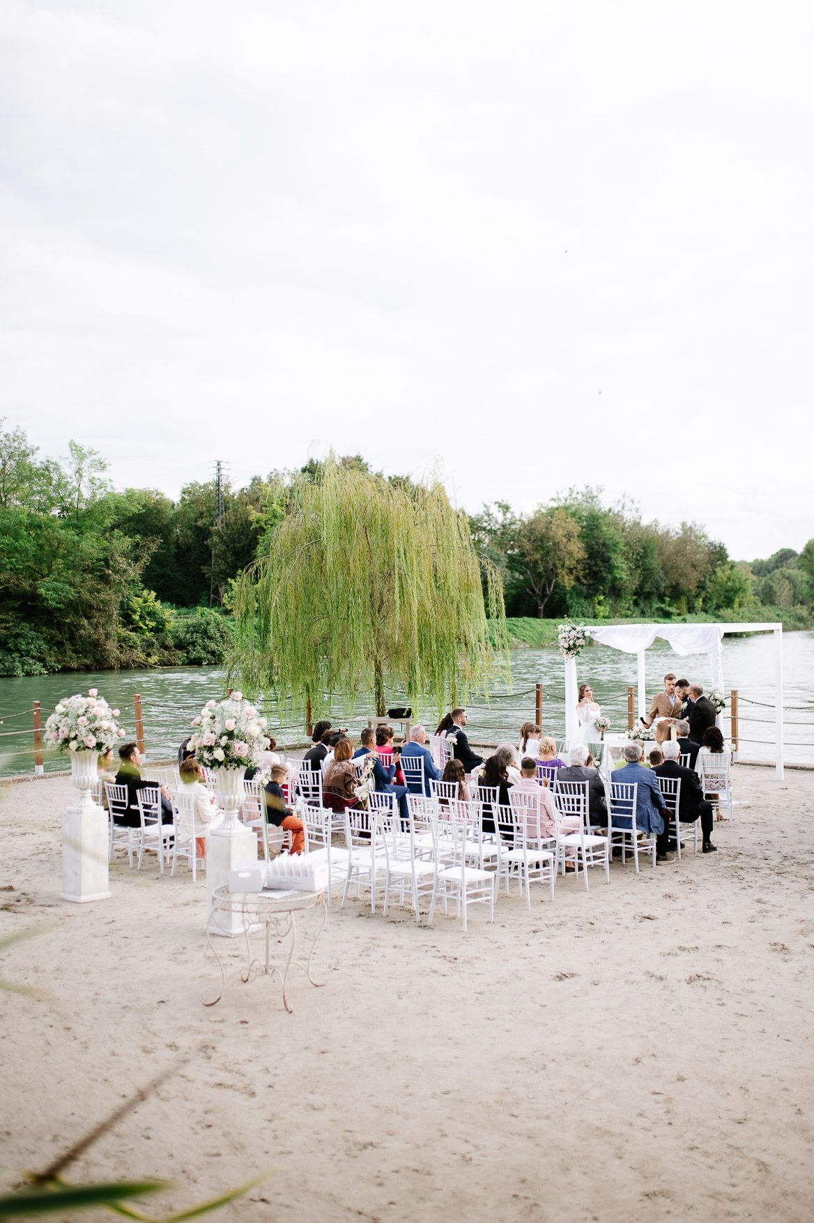 Beach wedding ceremony setup with white chairs overlooking a scenic waterfront with weeping willows.