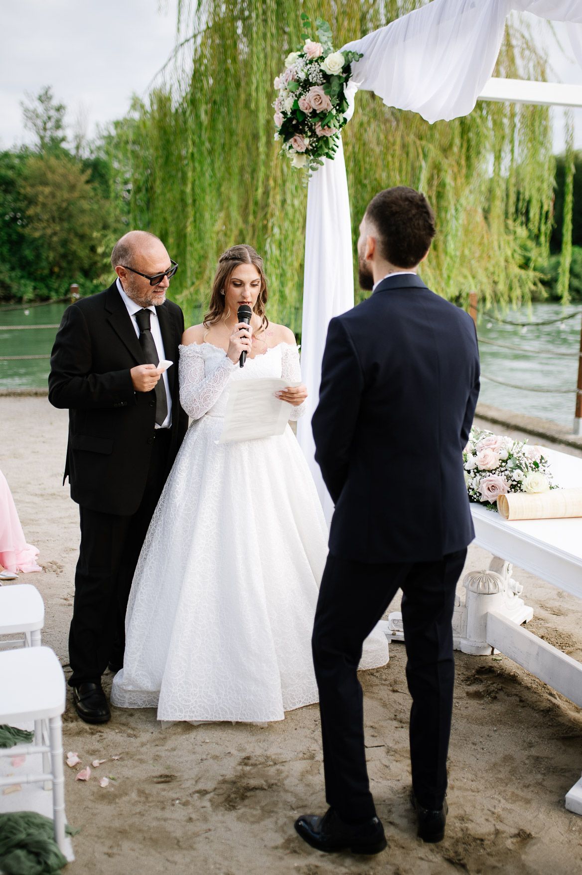 Wedding ceremony under a willow tree with bride in white dress reading vows beside floral arch decor.