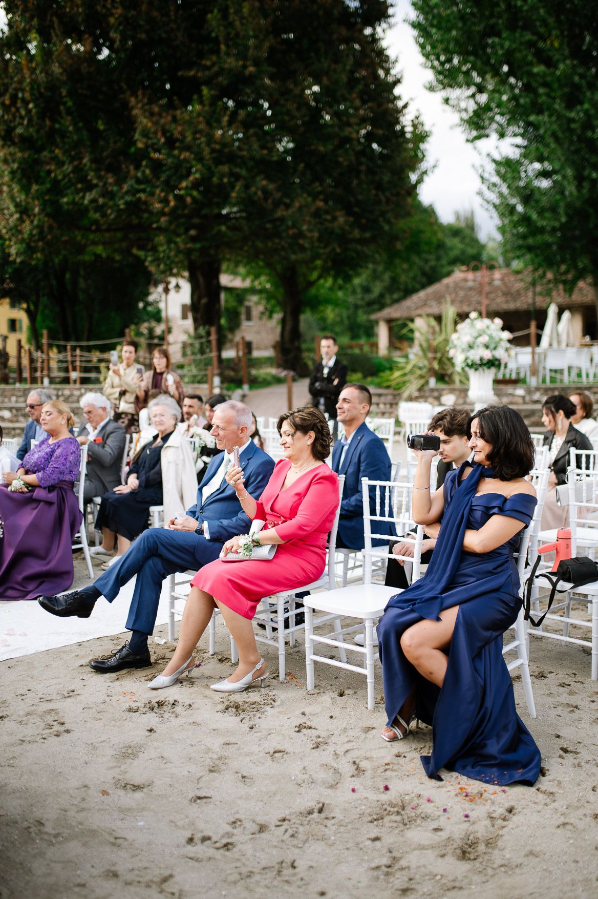 Wedding guests seated on white chairs on sandy beach during outdoor ceremony.