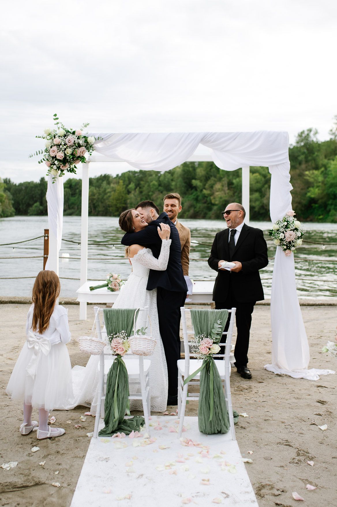 A waterfront wedding ceremony under a white draped arch with floral arrangements and rose petals on the aisle.