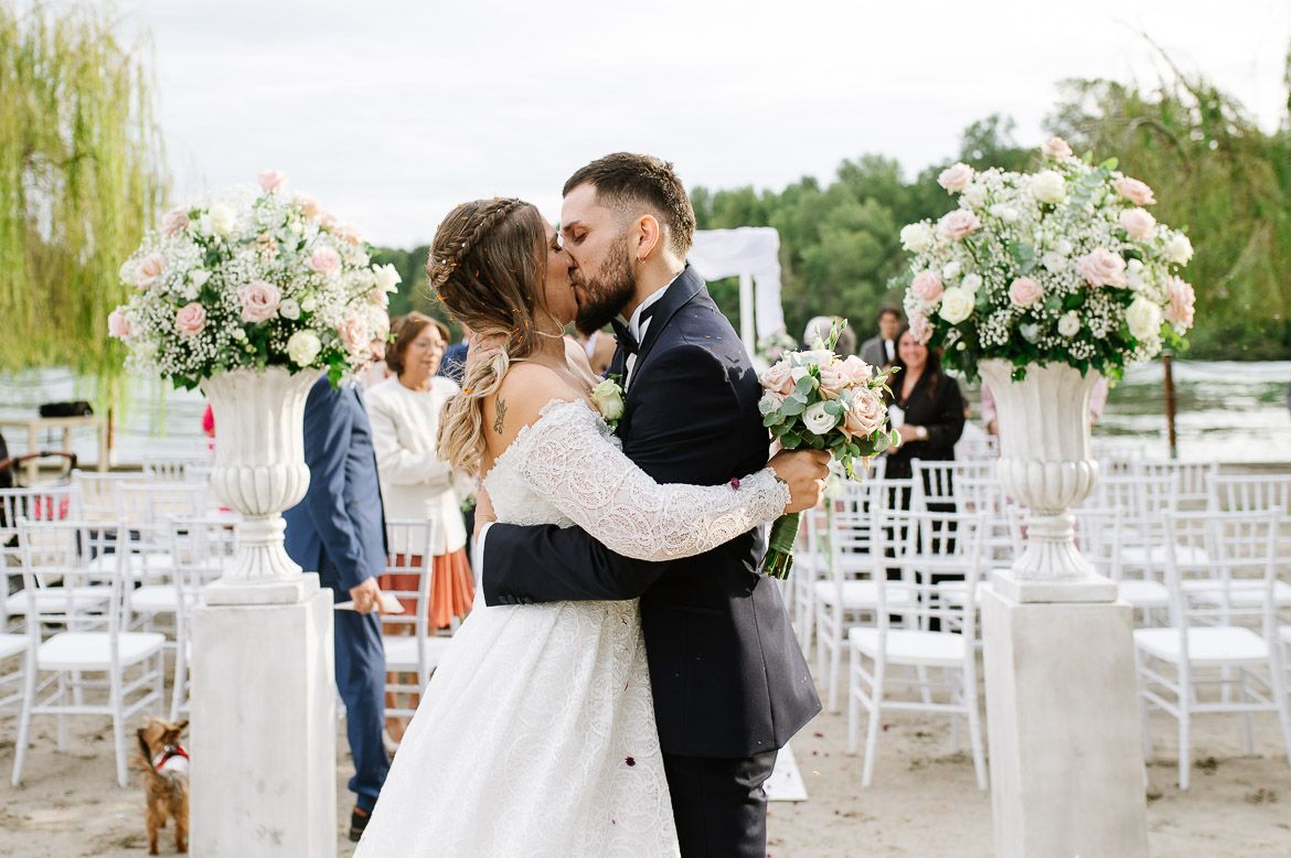 Outdoor wedding ceremony with white chairs and floral arrangements under weeping willow trees at a lakeside venue.