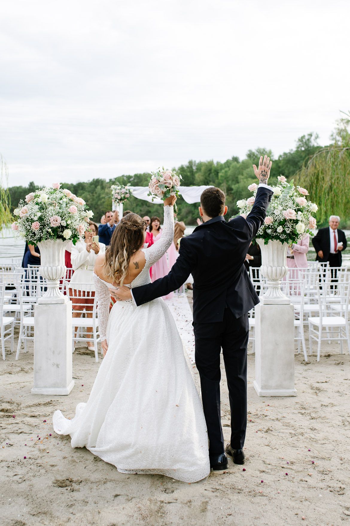 Wedding guests wave goodbye during beach ceremony exit with white chairs and floral arrangements.