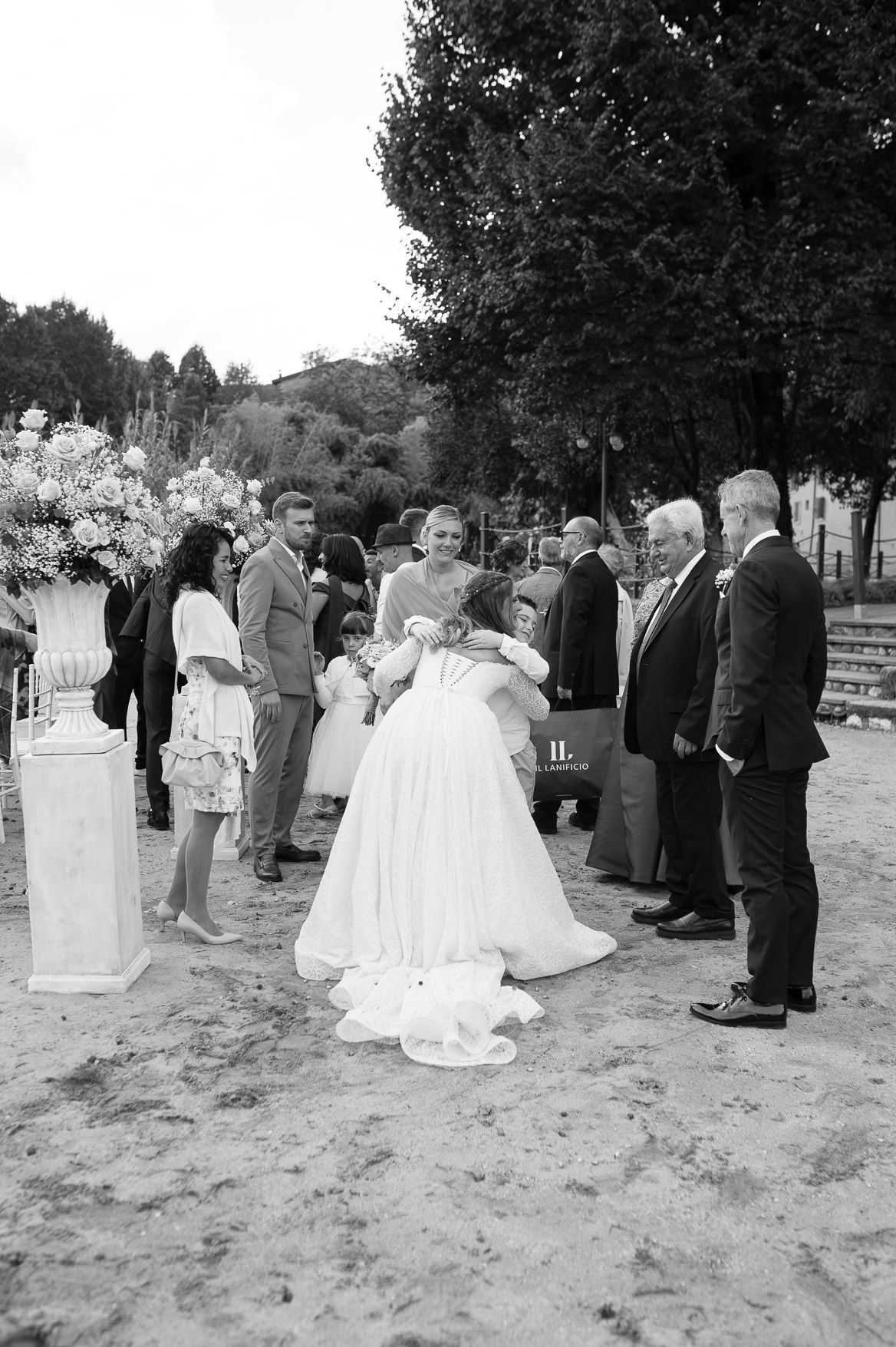 Black and white photo of wedding guests gathered outdoors on a sandy area during a celebration.