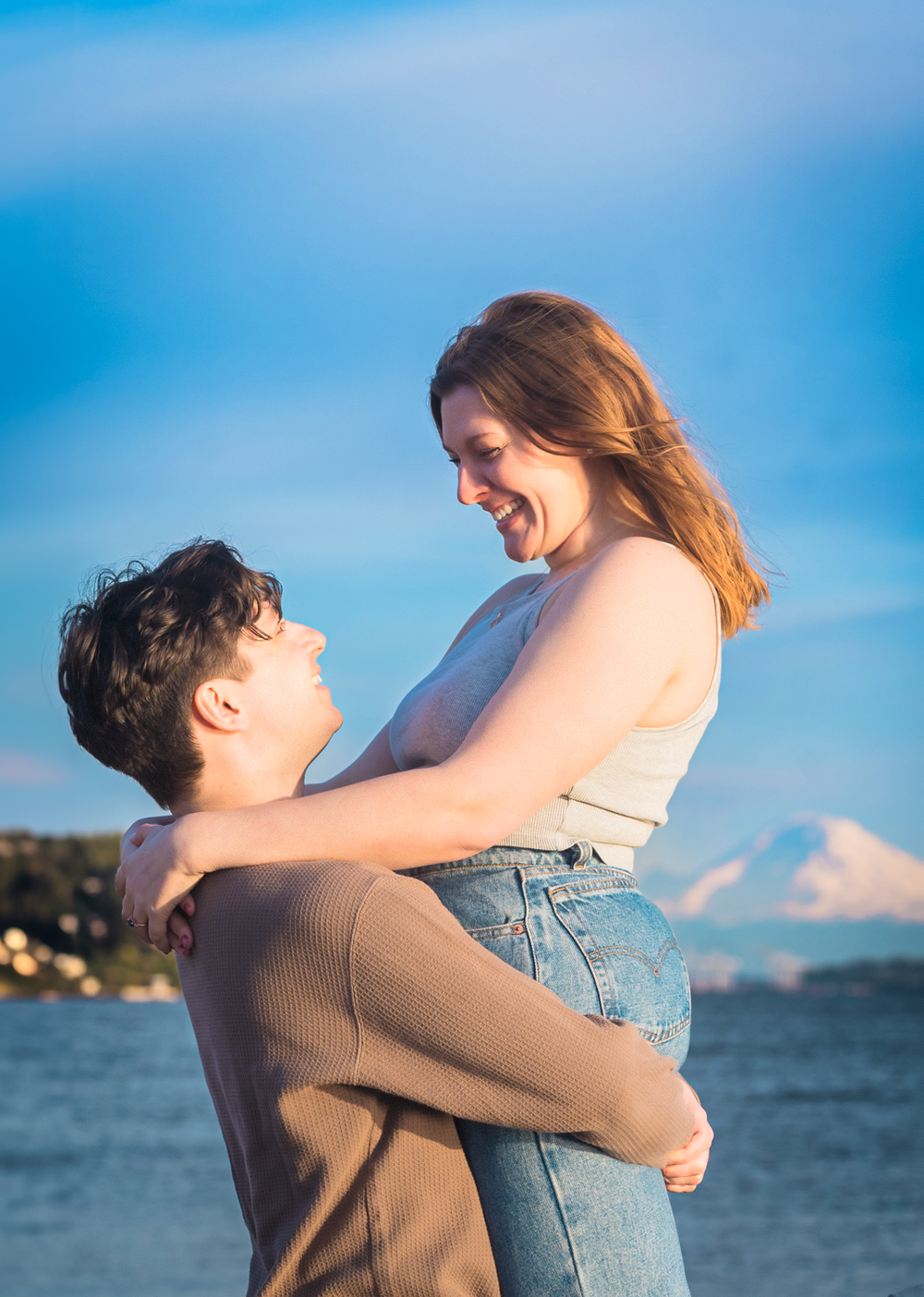 A couple embraces by the waterfront against a bright blue sky as the sun sets over the ocean.