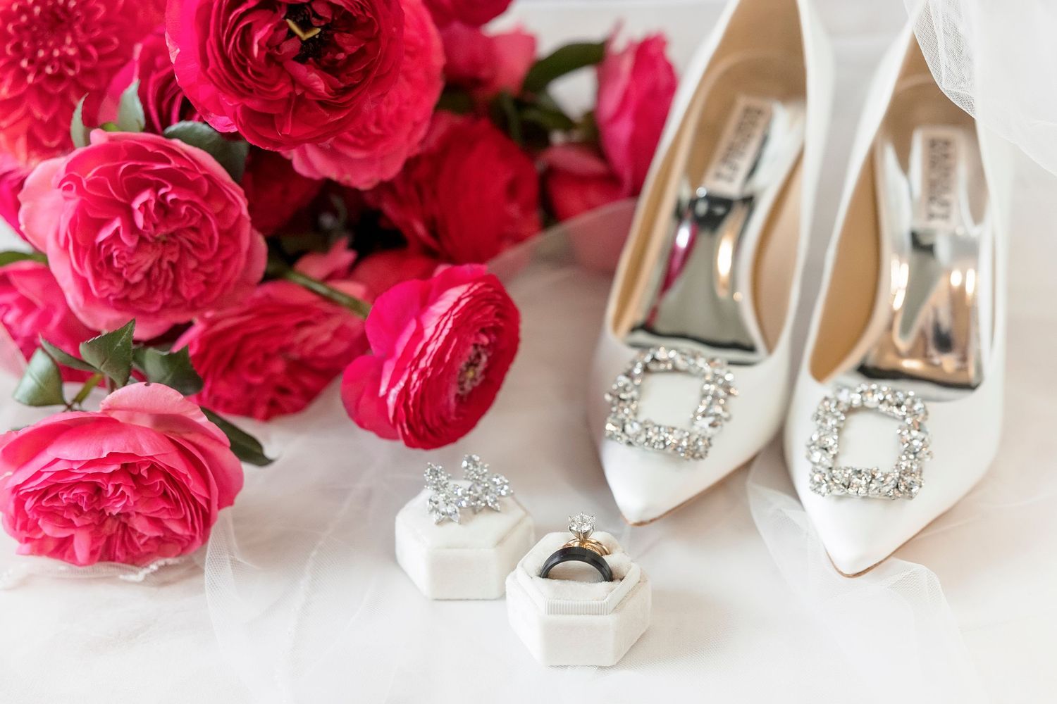 Elegant white bridal shoes with crystal embellishments displayed alongside vibrant pink carnation flowers.